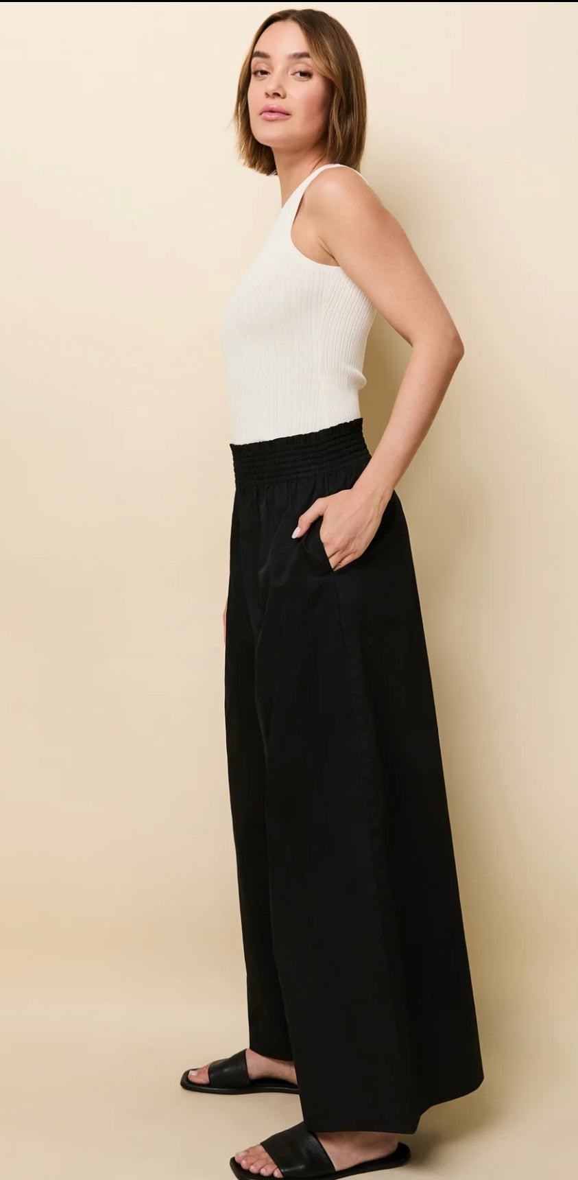 Woman wearing black high-waisted elastic waist Twyla Pant Noir palazzo pants by SOLID&STRIPED with white sleeveless top and black sandals