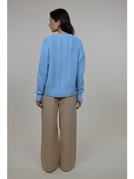 Back view of a woman wearing a light blue Cora Sweater with cable knit design and beige wide-leg pants against a neutral background