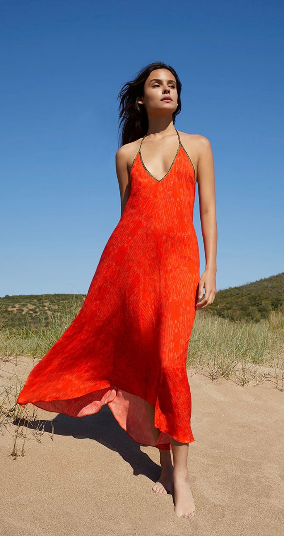 Woman wearing flowing Balinese Fire Red Rhodes Dress with low cut back standing on sandy beach under clear blue sky