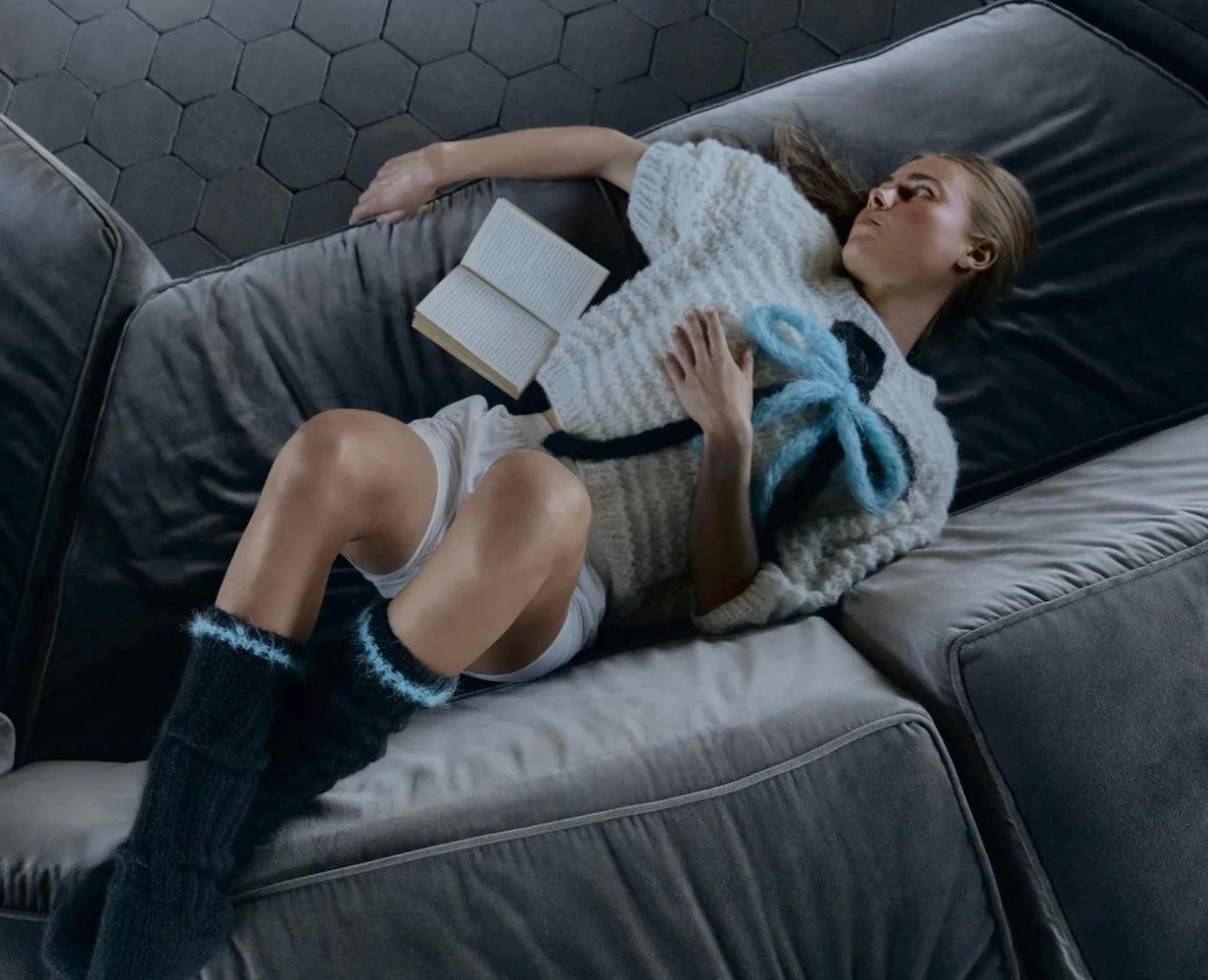Woman wearing cream Alpaca Puffy Top with knitted pattern lying on gray couch reading a book