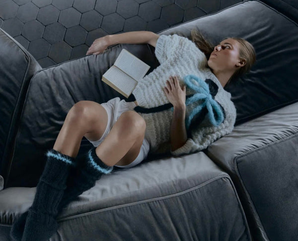 Woman reclining on gray couch wearing crème Alpaca Puffy Top with blue bow and matching dark knit socks