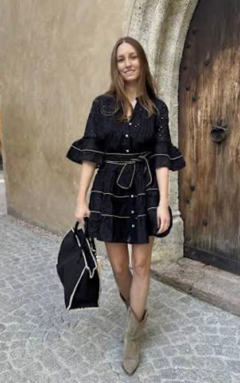 Woman wearing The Monte Carlo limited edition black dress with a belt, holding a matching black bag and standing on a cobblestone street.