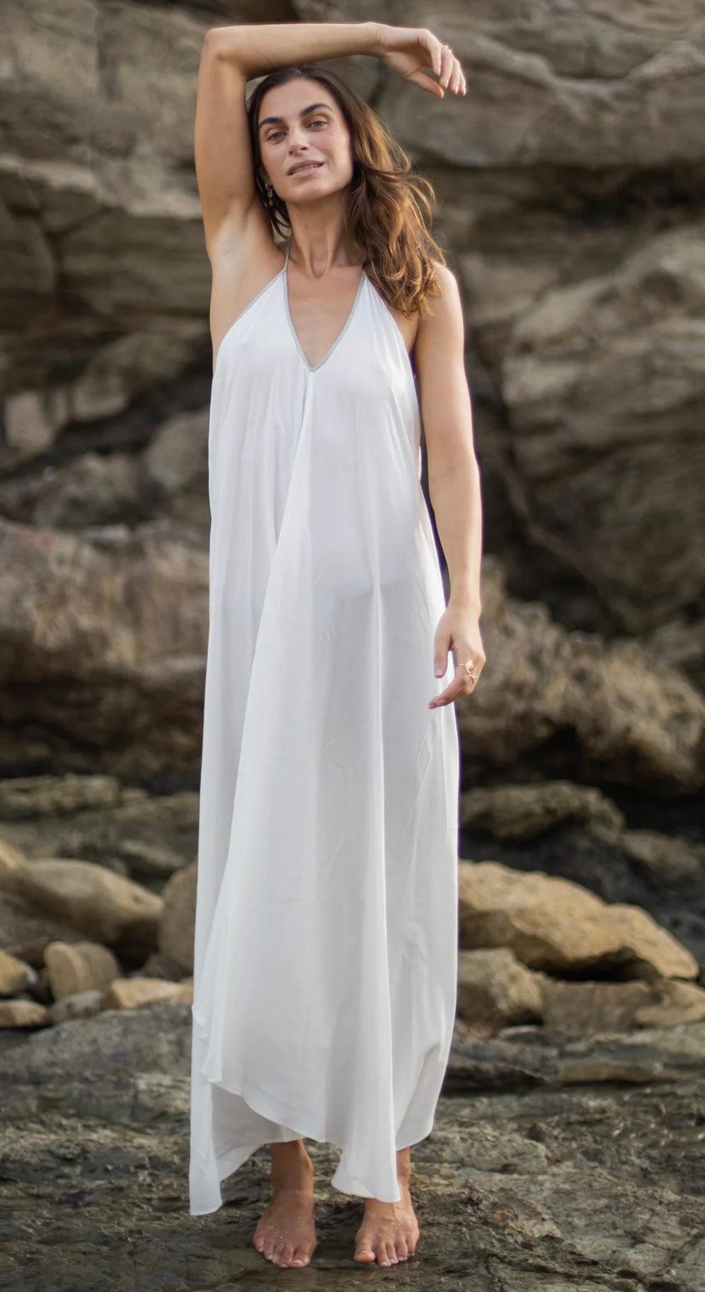 Woman wearing white Rhodes Dress with plunging V-neck and flowy high low hem standing on rocky beach