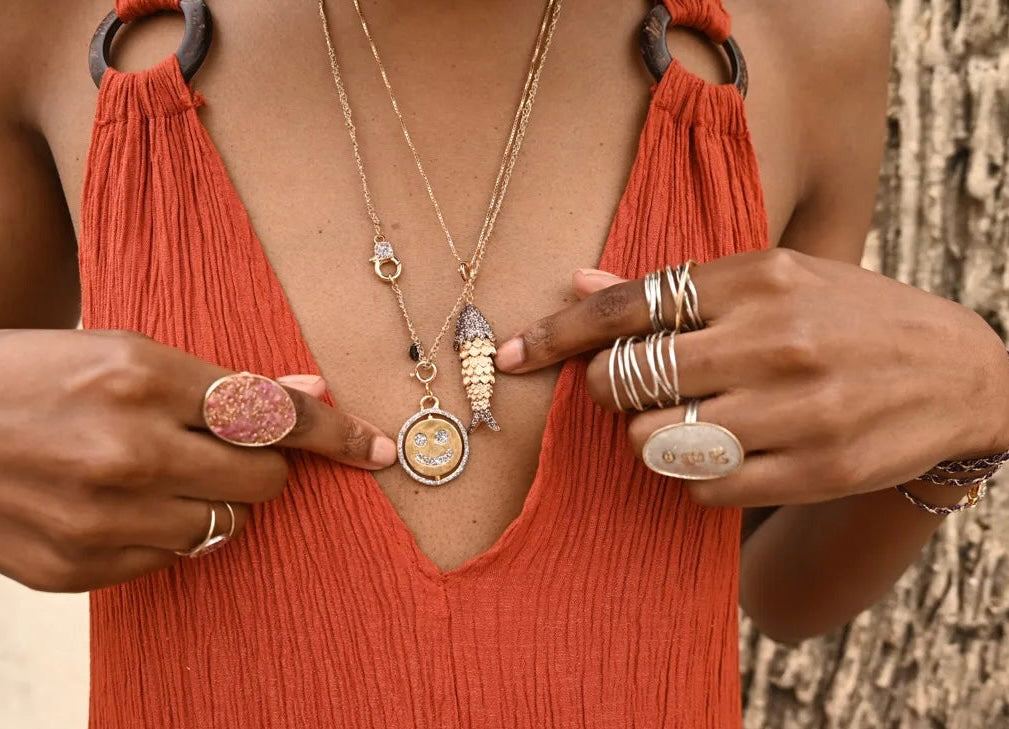 Close-up of a person wearing the Loulou Lucky Fish Necklace 18k gold plated with fish and coin pendants, styled with multiple rings and an orange textured top