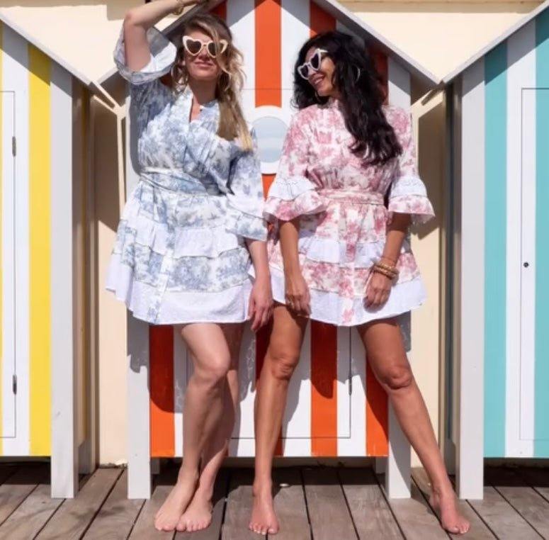 Two women wearing The Monte Carlo limited edition signature dresses standing barefoot in front of colorful striped beach huts